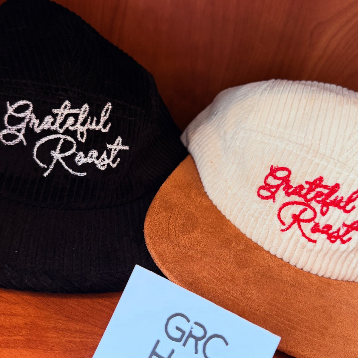 Two corduroy baseball caps with 'Grateful Roast' embroidered on the front. One cap is black with white embroidery and a black visor, and the other is cream with a tan visor and red embroidery.